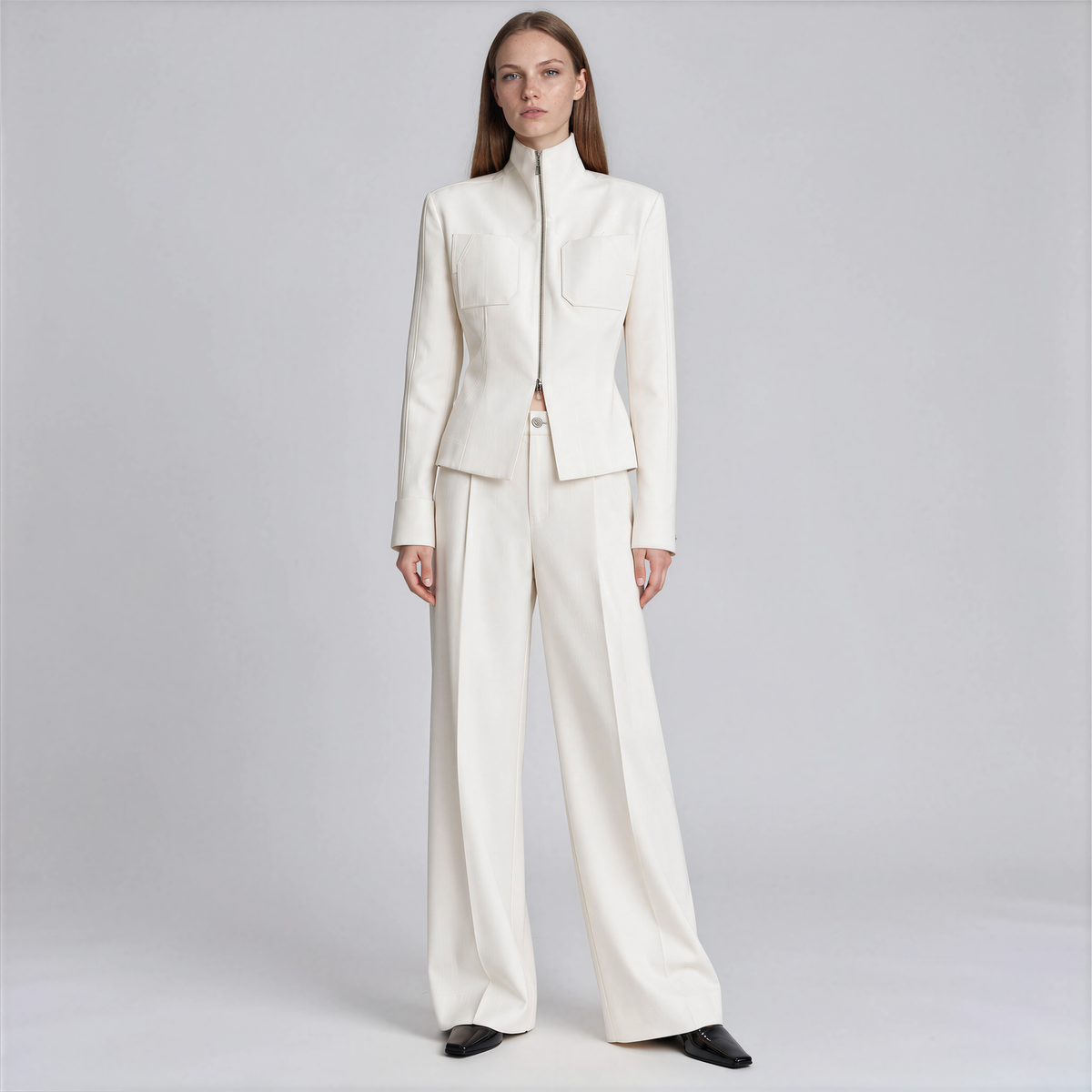 A model stands against a light gray studio background wearing a structured cream-colored two-piece suit consisting of a high-neck jacket and matching wide-leg trousers.