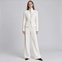 A model stands against a light gray studio background wearing a structured cream-colored two-piece suit consisting of a high-neck jacket and matching wide-leg trousers.