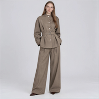 A woman modeling a taupe-brown two-piece linen set consisting of a belted button-up jacket and matching wide-leg trousers, shot against a light gray studio background.
