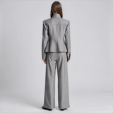 A model viewed from the back wearing a light gray two-piece suit with a structured blazer and wide-leg trousers, set against a plain light gray studio background.