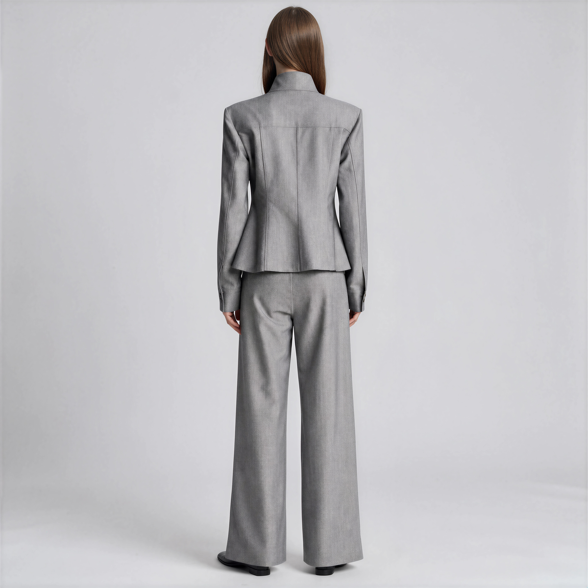 A model viewed from the back wearing a light gray two-piece suit with a structured blazer and wide-leg trousers, set against a plain light gray studio background.