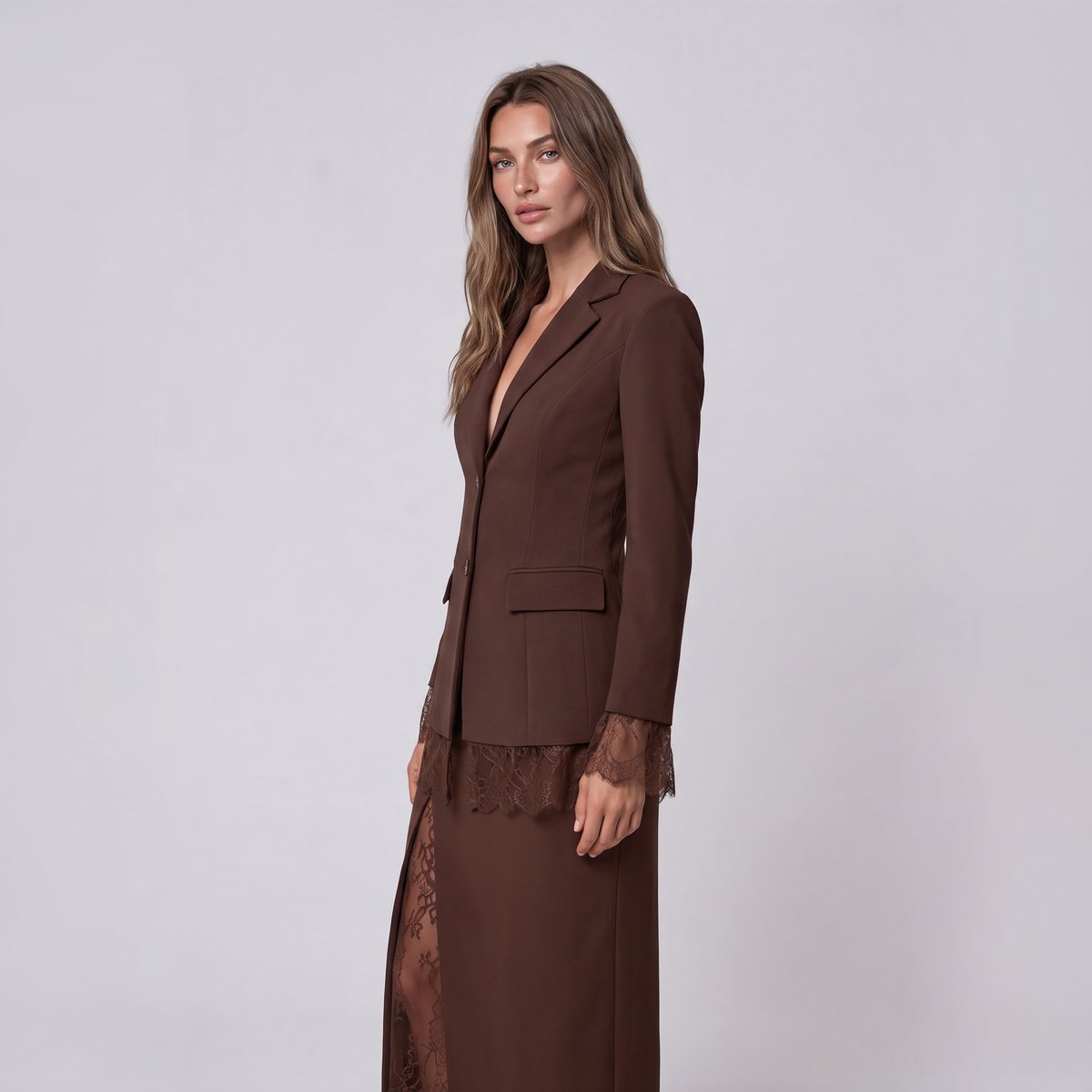 A woman modeling a dark brown tailored blazer with matching long skirt, featuring sheer lace trim at the hem and cuffs, against a plain light gray studio background.