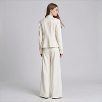 A woman stands with her back to the camera wearing a matching cream-colored tailored suit consisting of a cropped blazer and wide-leg trousers, set against a plain light gray studio background.