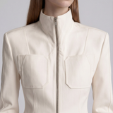 A close-up of a model wearing a cream-colored structured jacket with a high stand-up collar, front zipper, and two large chest pockets.