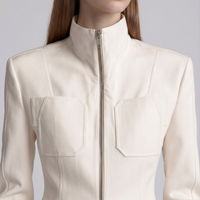 A close-up of a model wearing a cream-colored structured jacket with a high stand-up collar, front zipper, and two large chest pockets.