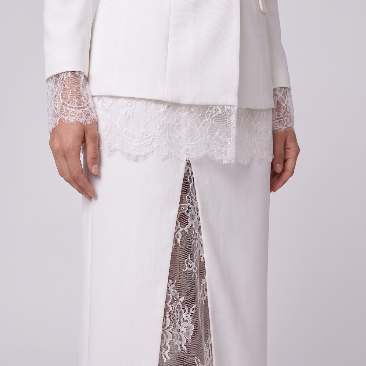 White tailored suit with sheer lace trim at the cuffs and waist, worn by a model. The white trousers feature a front slit revealing a sheer white lace insert panel.