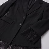 A close-up of a black blazer with notched lapels, two dark buttons, flap pockets, and black lace trim at the cuffs and hem, laid flat on a light gray background.
