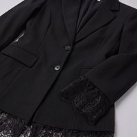 A close-up of a black blazer with notched lapels, two dark buttons, flap pockets, and black lace trim at the cuffs and hem, laid flat on a light gray background.