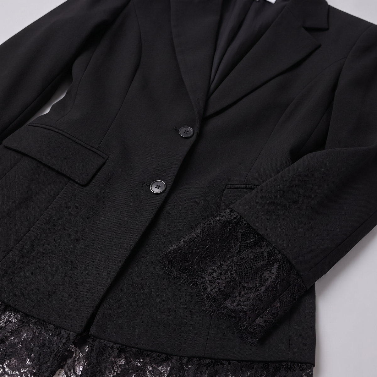 A close-up of a black blazer with notched lapels, two dark buttons, flap pockets, and black lace trim at the cuffs and hem, laid flat on a light gray background.