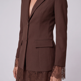 A close-up of a model wearing a dark brown tailored blazer with notched lapels, a single front button, and a flap pocket, paired with a matching lace-trimmed skirt against a light gray studio background.