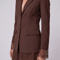 A close-up of a model wearing a dark brown tailored blazer with notched lapels, a single front button, and a flap pocket, paired with a matching lace-trimmed skirt against a light gray studio background.