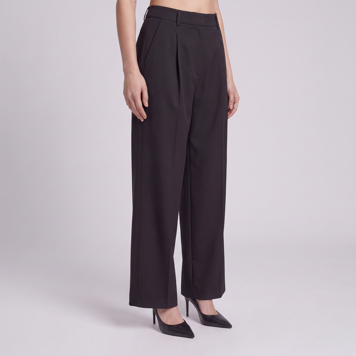A model wearing black wide-leg pleated trousers and black pointed-toe heels, shown from the waist down against a light gray studio background.