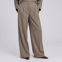 A person wearing high-waisted, wide-leg taupe trousers with pleats and a matching belt, paired with dark brown pointed-toe shoes, photographed against a light gray studio background.