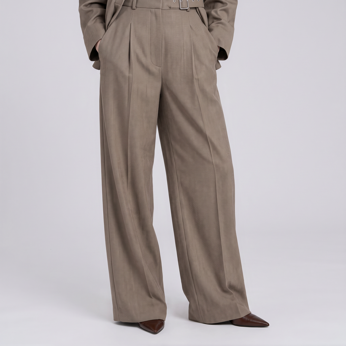 A person wearing high-waisted, wide-leg taupe trousers with pleats and a matching belt, paired with dark brown pointed-toe shoes, photographed against a light gray studio background.