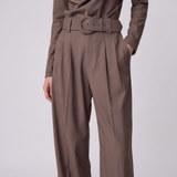 A person wearing high-waisted taupe-brown pleated trousers with a matching belt, paired with a long-sleeve top, against a light gray studio background.