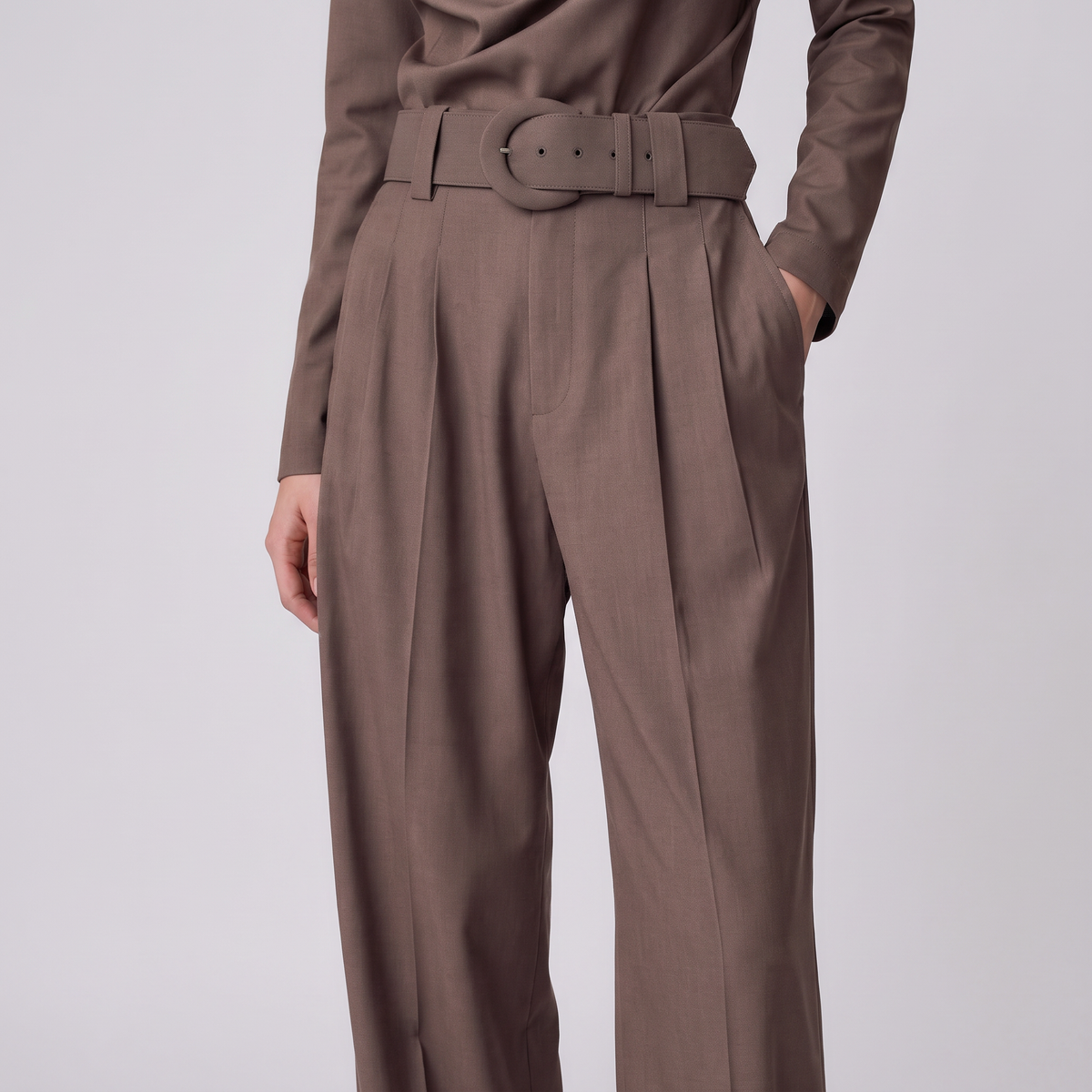 A person wearing high-waisted taupe-brown pleated trousers with a matching belt, paired with a long-sleeve top, against a light gray studio background.