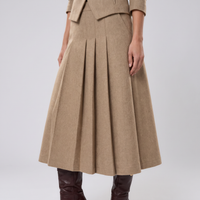 A model wearing a high-waisted, pleated A-line skirt in a warm camel beige color, paired with dark brown leather boots, shown from the waist down against a light gray studio background.