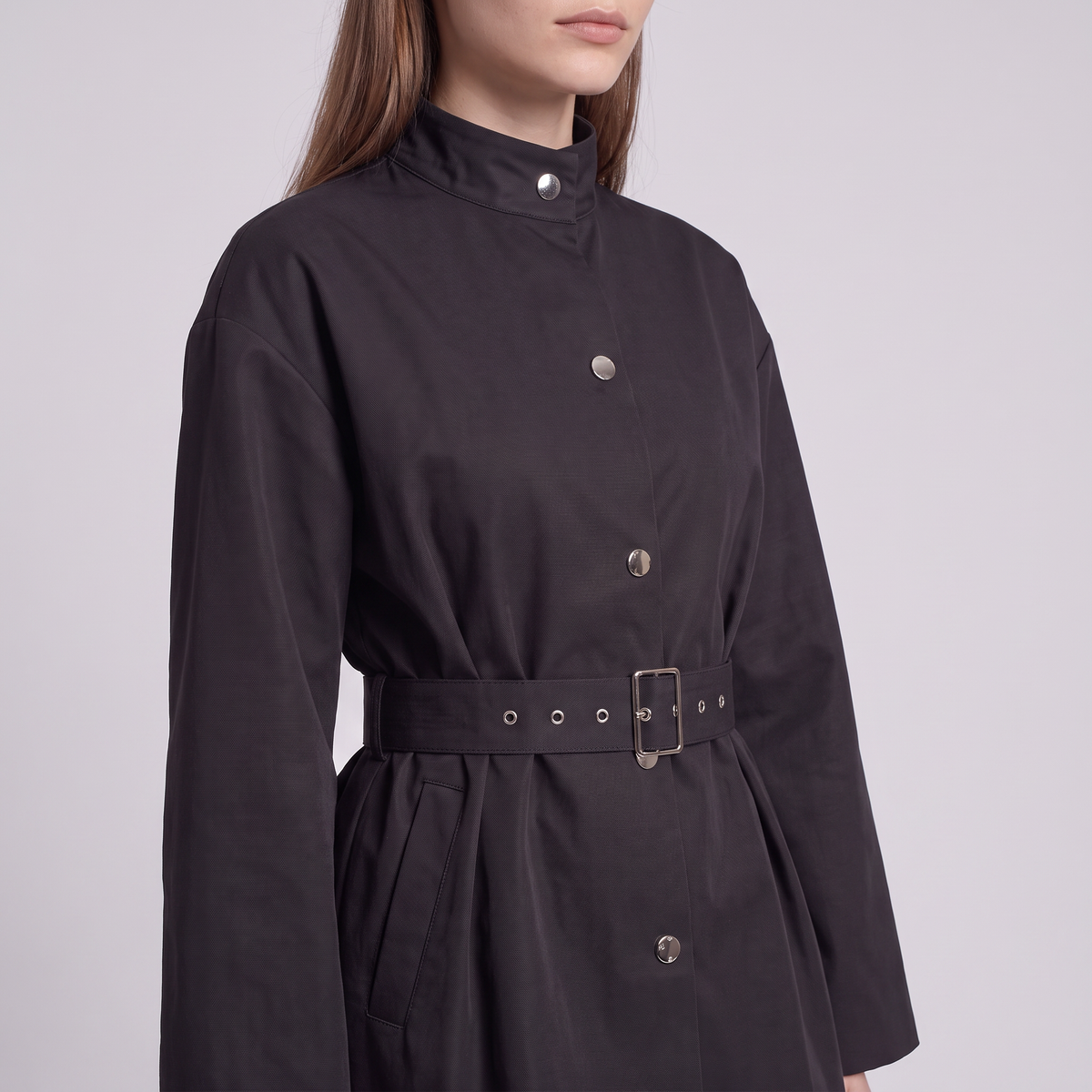 A close-up of a model wearing a black long-sleeve high-neck coat with a matching waist belt and silver-tone buttons, set against a plain light gray studio background.