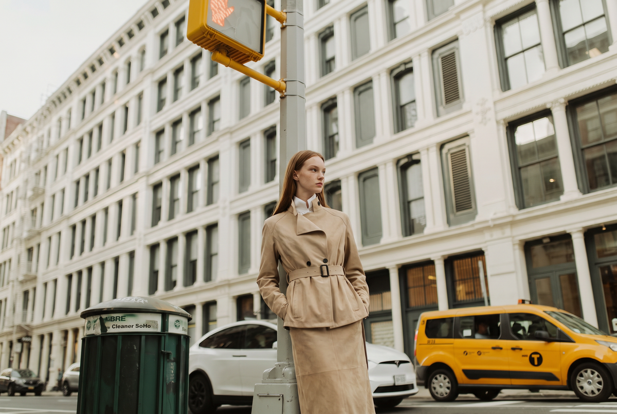 A woman wearing a beige belted trench coat and matching midi skirt stands on a city street in front of a large white building in New York City, with a yellow cab and street elements in the background