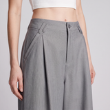 A close-up of a model wearing high-waisted, wide-leg gray trousers with pleated detailing and belt loops, paired with a white cropped top against a light gray studio background.