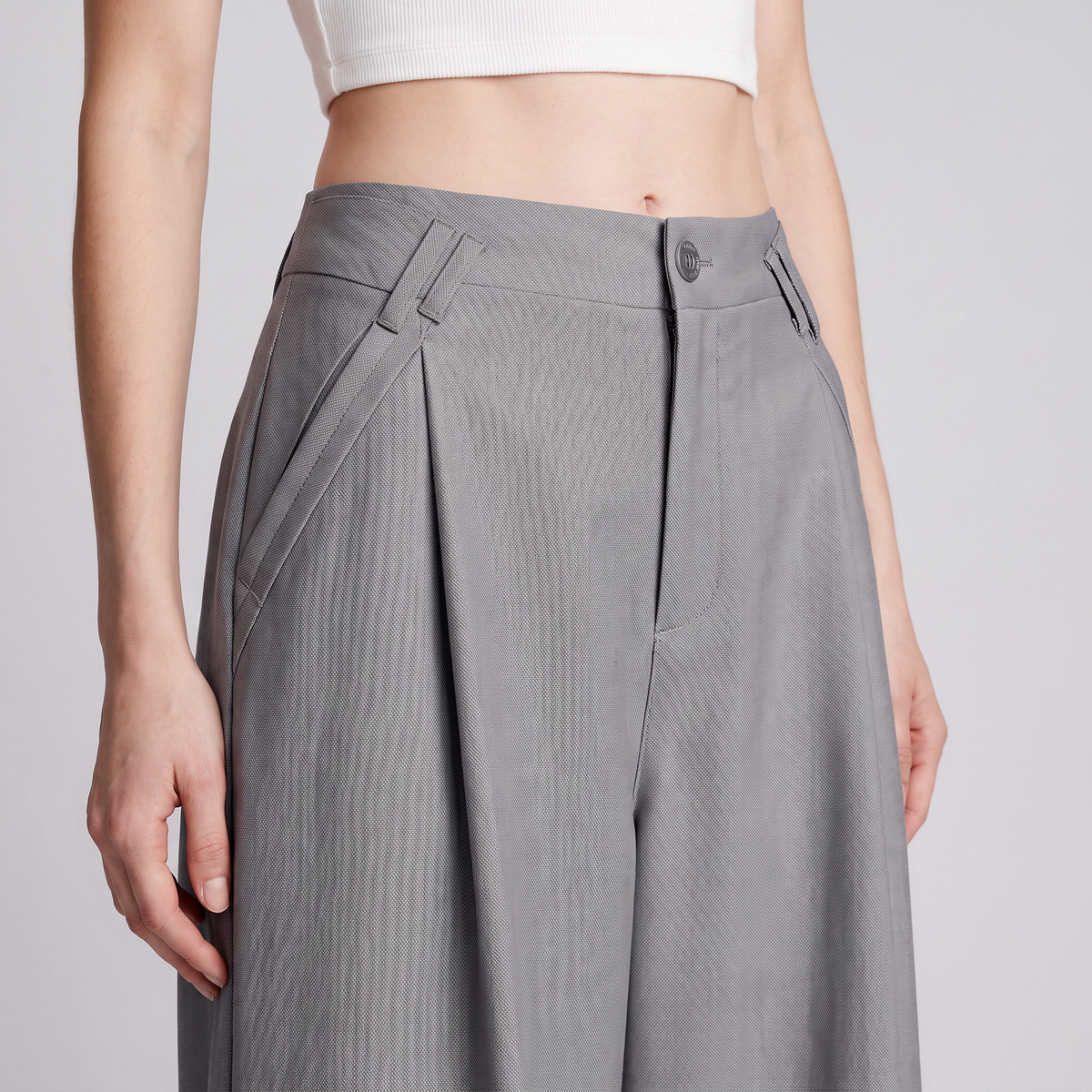 A close-up of a model wearing high-waisted, wide-leg gray trousers with pleated detailing and belt loops, paired with a white cropped top against a light gray studio background.