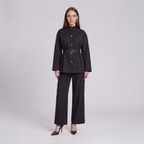 A model stands against a light gray studio background wearing a black long-sleeve belted jacket with a high mock neck and matching wide-leg trousers.
