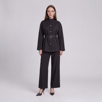 A model stands against a light gray studio background wearing a black long-sleeve belted jacket with a high mock neck and matching wide-leg trousers.
