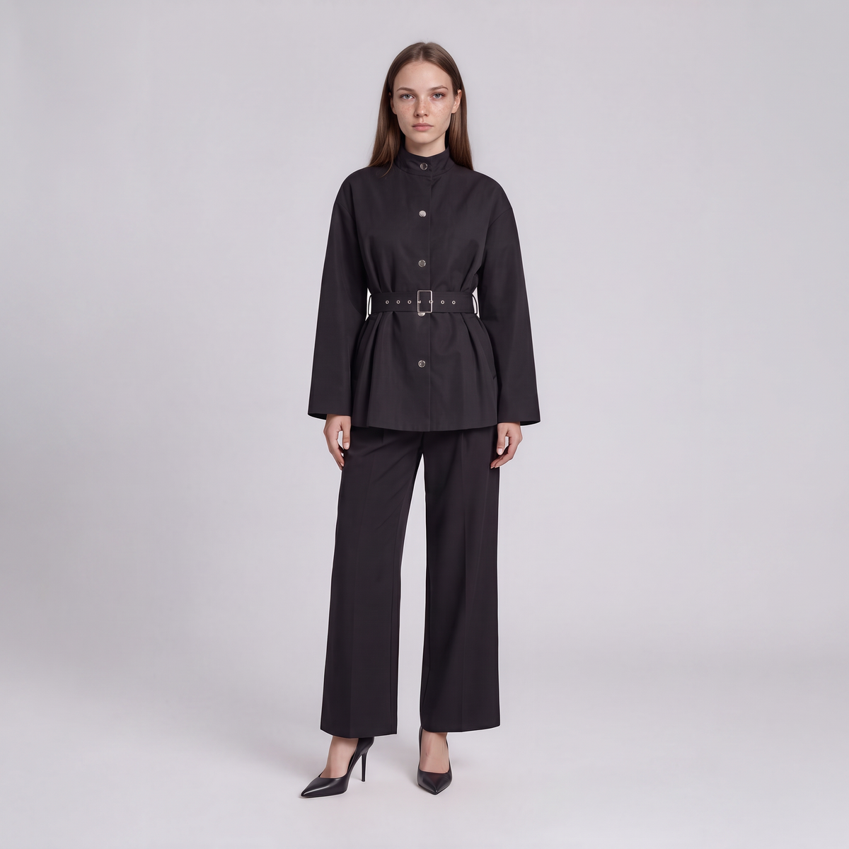 A model stands against a light gray studio background wearing a black long-sleeve belted jacket with a high mock neck and matching wide-leg trousers.