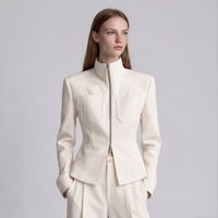 A woman modeling a structured cream-colored jacket with a high stand-up collar, front zipper, and flap chest pockets, paired with matching trousers against a light gray studio background.