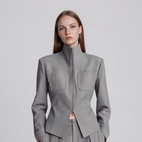 A woman modeling a structured light gray jacket with a high stand collar, front zipper, and two large chest pockets, photographed against a plain light gray studio background.