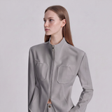 A woman wearing a structured light gray jacket with a stand collar, front zipper, and two large chest pockets, photographed against a plain light gray studio background.