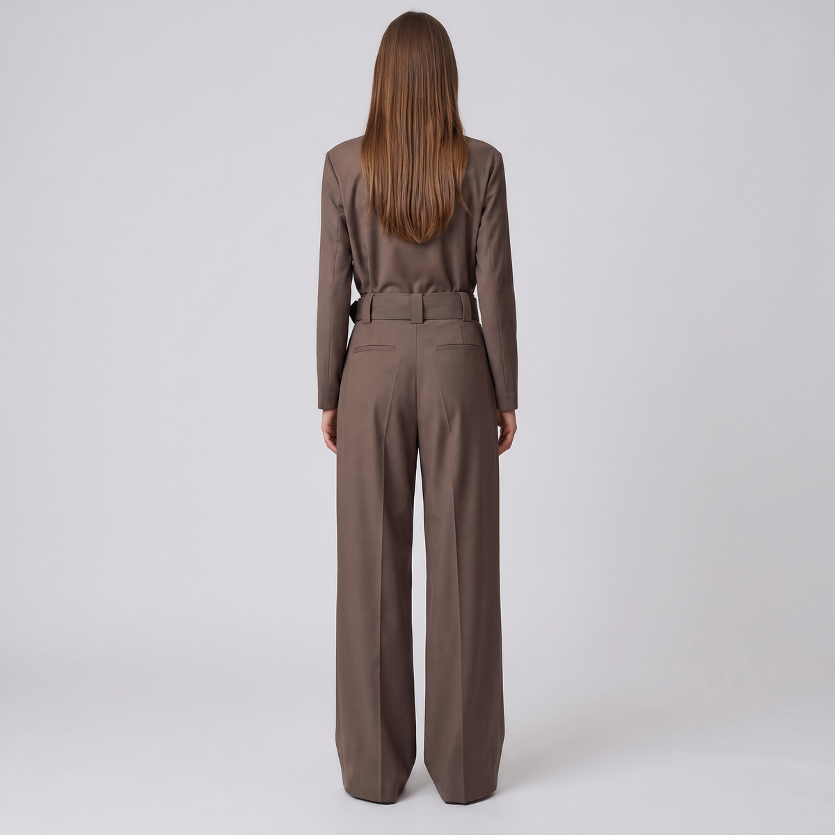 A woman viewed from behind wearing a matching brown long-sleeve top and wide-leg trousers set with a matching belt, against a plain light gray studio background.