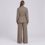 A model viewed from the back wearing a taupe-brown belted blazer and matching wide-leg trousers against a light gray studio background.