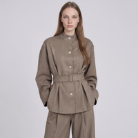 A woman modeling a taupe-brown belted jacket with a matching pair of trousers against a light gray studio background.