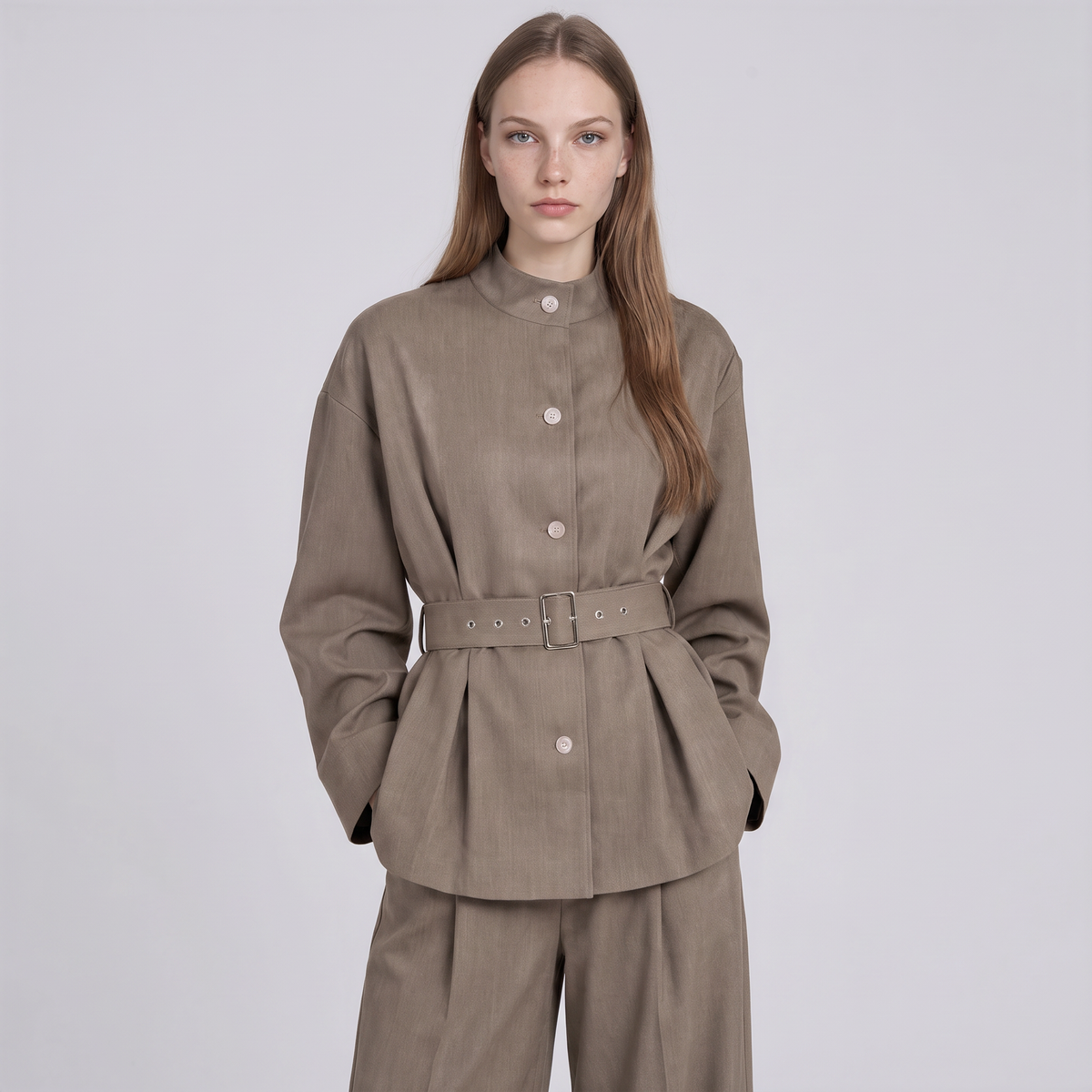 A woman modeling a taupe-brown belted jacket with a matching pair of trousers against a light gray studio background.