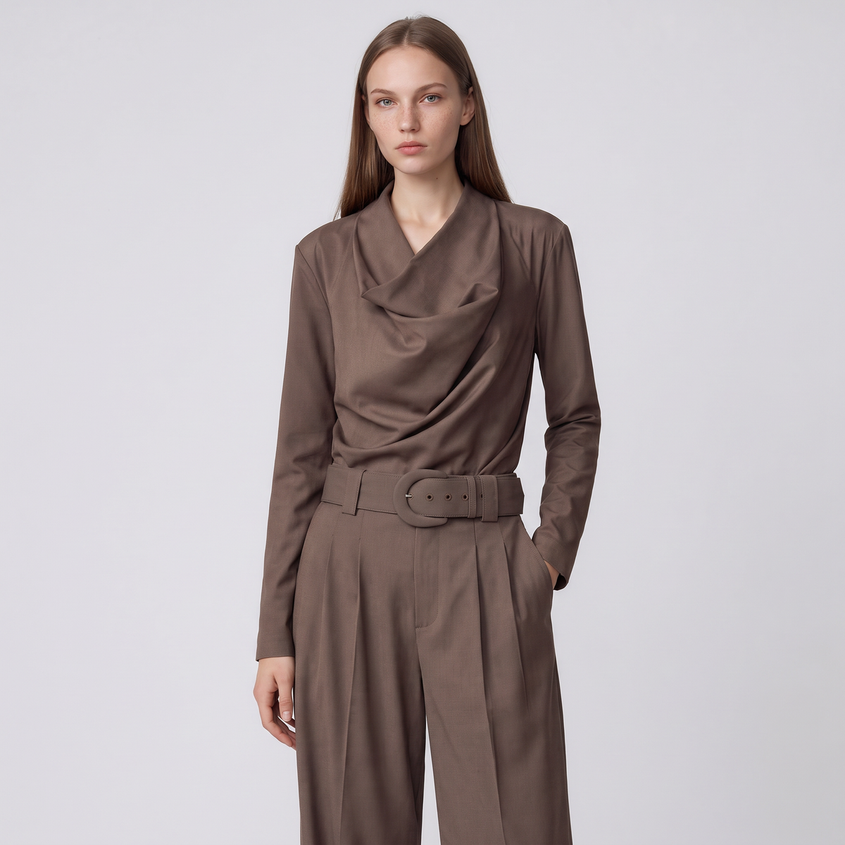 A woman modeling a brown draped long-sleeve top and matching wide-leg trousers with a matching belt, set against a plain light gray studio background.