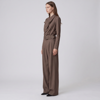 A woman stands in profile wearing a taupe-brown long-sleeve jumpsuit with a draped neckline, a matching belt, and wide-leg trousers, photographed against a light gray studio background.