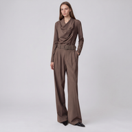 A woman models a brown long-sleeve jumpsuit with a draped cowl neckline, a matching belt, and wide-leg trousers, set against a plain light gray studio background.