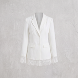 A white tailored blazer with notched lapels, two front buttons, flap pockets, and lace trim at the cuffs and hem, displayed on a mannequin against a light textured wall.