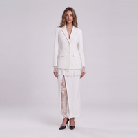 A woman stands against a light gray studio background wearing a white tailored blazer with lace cuffs, a white skirt, and a sheer white lace overlay panel, paired with black pointed heels.