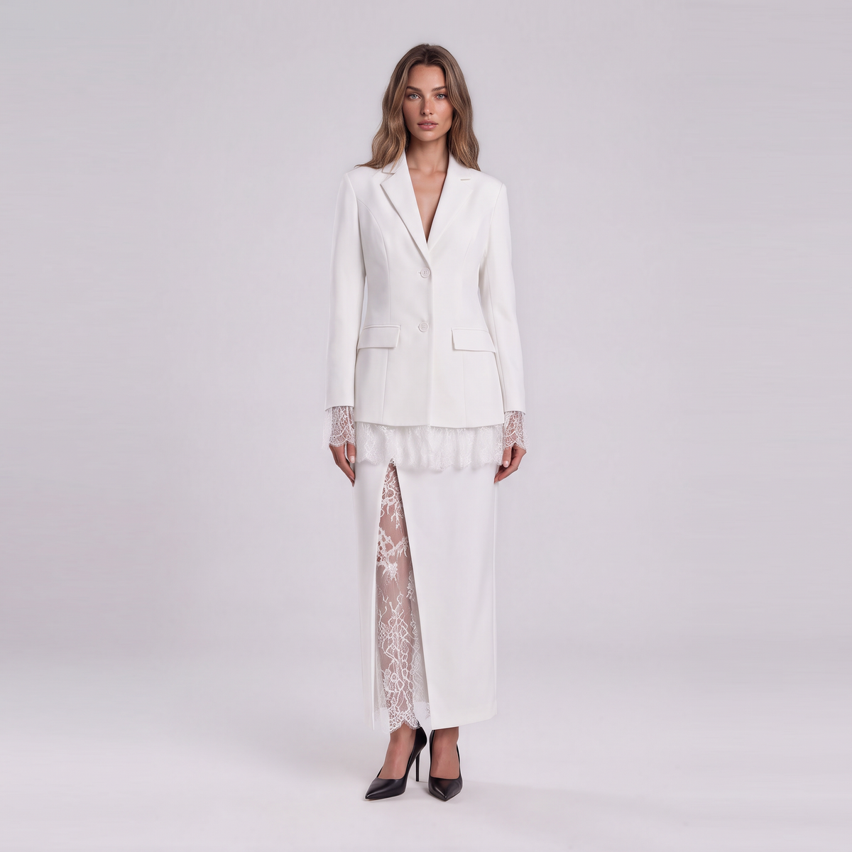 A woman stands against a light gray studio background wearing a white tailored blazer with lace cuffs, a white skirt, and a sheer white lace overlay panel, paired with black pointed heels.