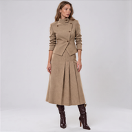 A woman modeling a beige double-breasted midi coat with a matching pleated skirt, paired with dark brown knee-high boots, against a light gray studio background.