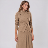 A woman modeling a matching tan two-piece outfit consisting of a structured long-sleeve jacket with a high mock neck and a matching pleated midi skirt, set against a plain light gray studio background.