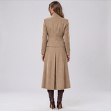 A woman viewed from the back wearing a camel-colored long coat with structured shoulders and a flared skirt, paired with dark brown knee-high boots, against a light gray studio background.