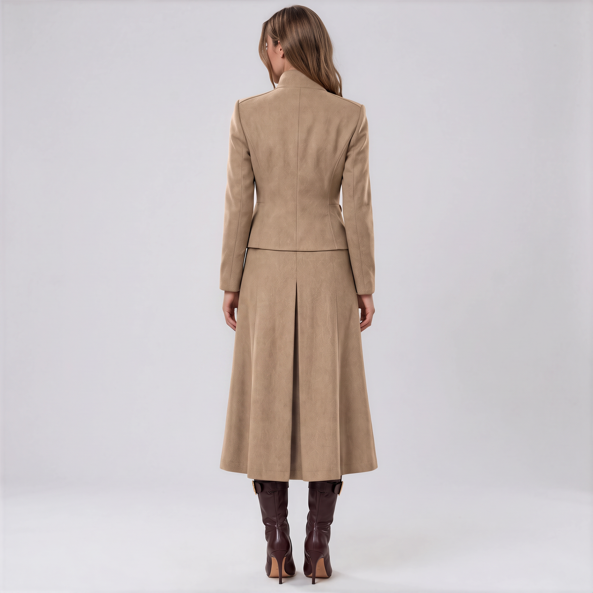 A woman viewed from the back wearing a camel-colored long coat with structured shoulders and a flared skirt, paired with dark brown knee-high boots, against a light gray studio background.