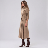 A woman models a beige two-piece suit consisting of a structured blazer with a high collar and matching pleated midi skirt, paired with dark brown ankle boots, against a light gray studio background.