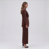 A woman stands in profile wearing a matching brown tailored set with a cropped blazer, wide-leg trousers, and fringe detail at the hem, holding a structured brown handbag against a light gray studio background.