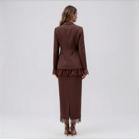 A woman stands with her back to the camera wearing a matching brown tailored jacket and long skirt set with lace trim, photographed against a plain light gray studio background.