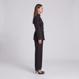 A woman stands in profile wearing a dark tailored two-piece outfit with a long-sleeve belted jacket and matching straight-leg pants, paired with black heels, against a light gray studio background.