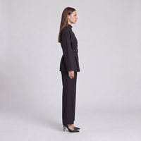 A woman stands in profile wearing a dark tailored two-piece outfit with a long-sleeve belted jacket and matching straight-leg pants, paired with black heels, against a light gray studio background.